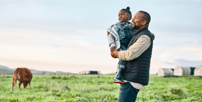 An adult is carrying a child while taking a walk on grassy a farm